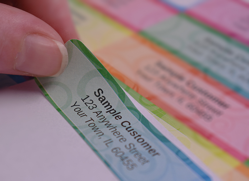 A close-up showing a hand peeling a personalized return address label from a sheet of colorful, patterned designs.